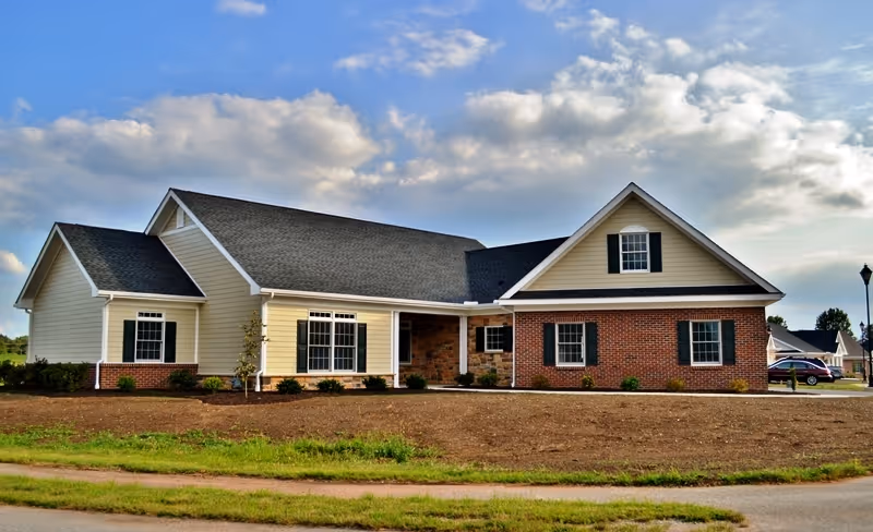 Single-story residential building with beige siding and red brick exterior, multiple windows with black shutters, and a gray shingled roof under a partly cloudy sky.