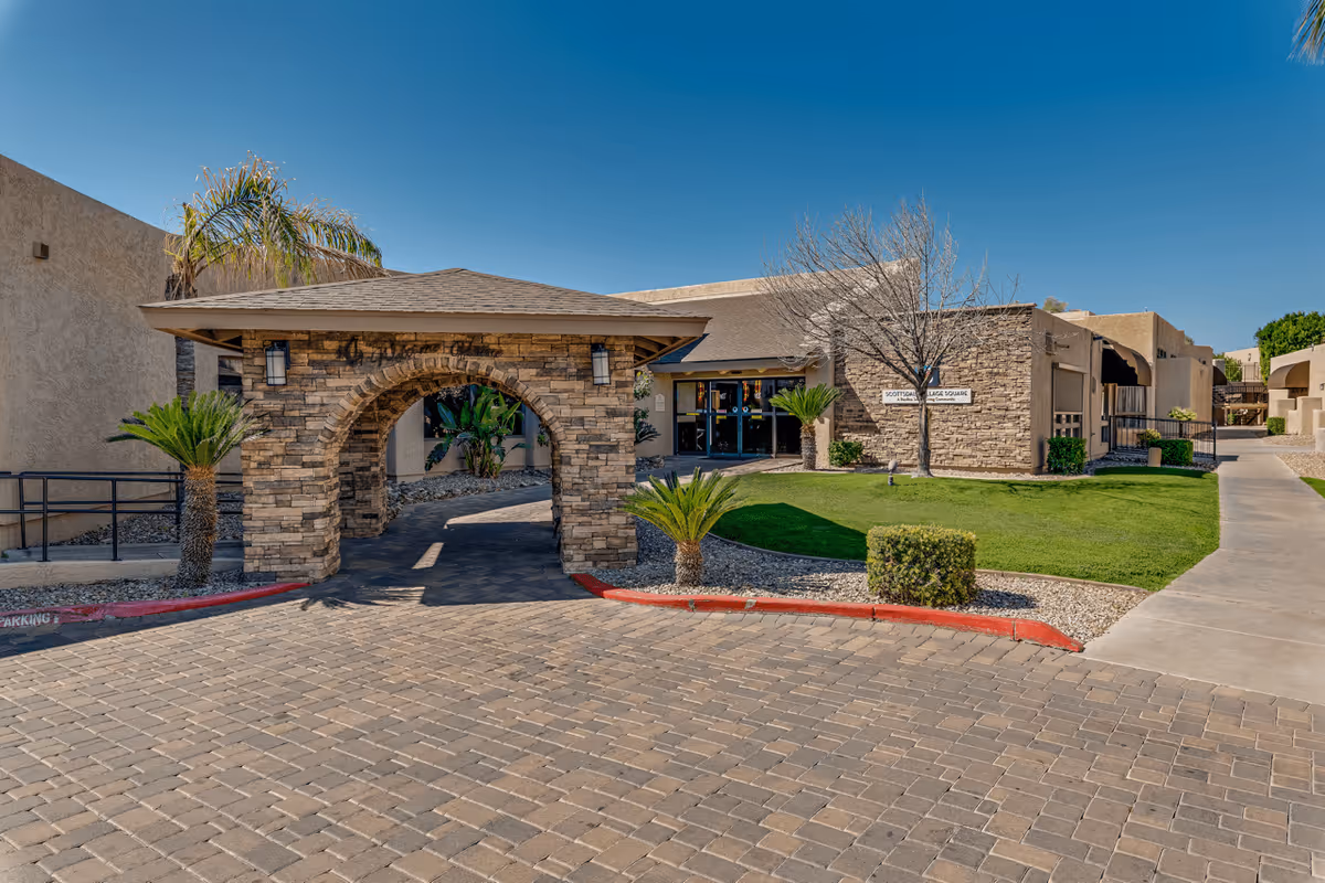 Exterior view of Scottsdale Village Square facility showing a stone archway entrance, paved driveway, green lawn, palm trees, and a clear blue sky.