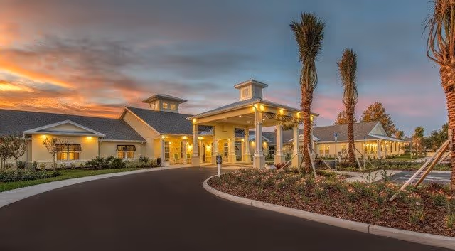 Exterior view of The Addison of Port Orange facility at sunset, showing a well-lit entrance with a covered drop-off area, palm trees, landscaped garden beds, and a curved driveway.
