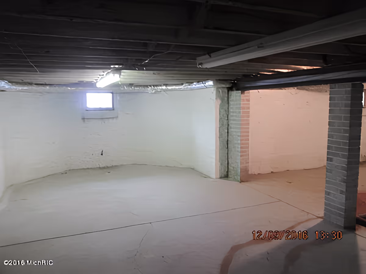 Empty basement room with white painted brick walls, a small window, exposed ceiling beams, and a concrete floor with visible cracks. A fluorescent light fixture is mounted on the ceiling, and there is a brick support column near the center-right.