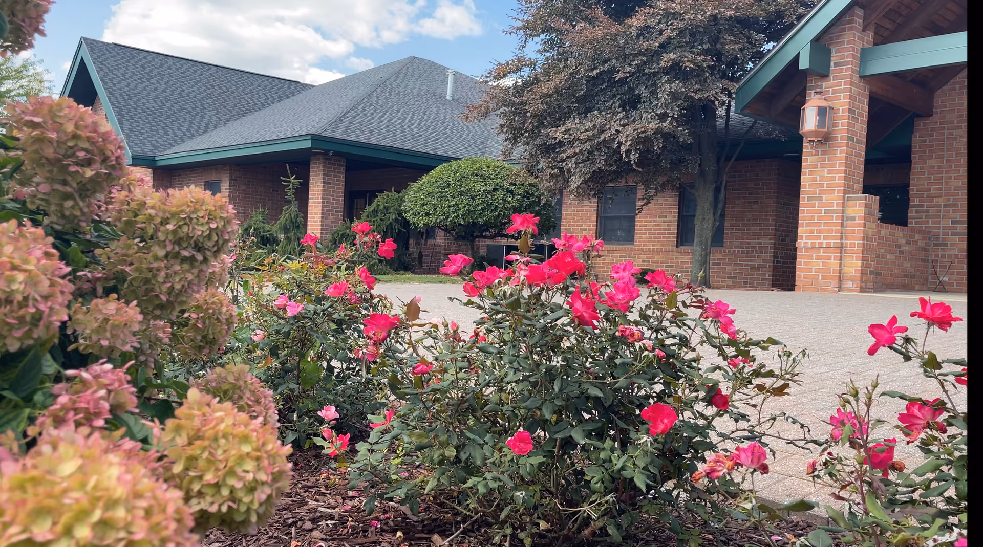 Brick building entrance with pink roses and hydrangeas in the foreground under a gabled roof.
