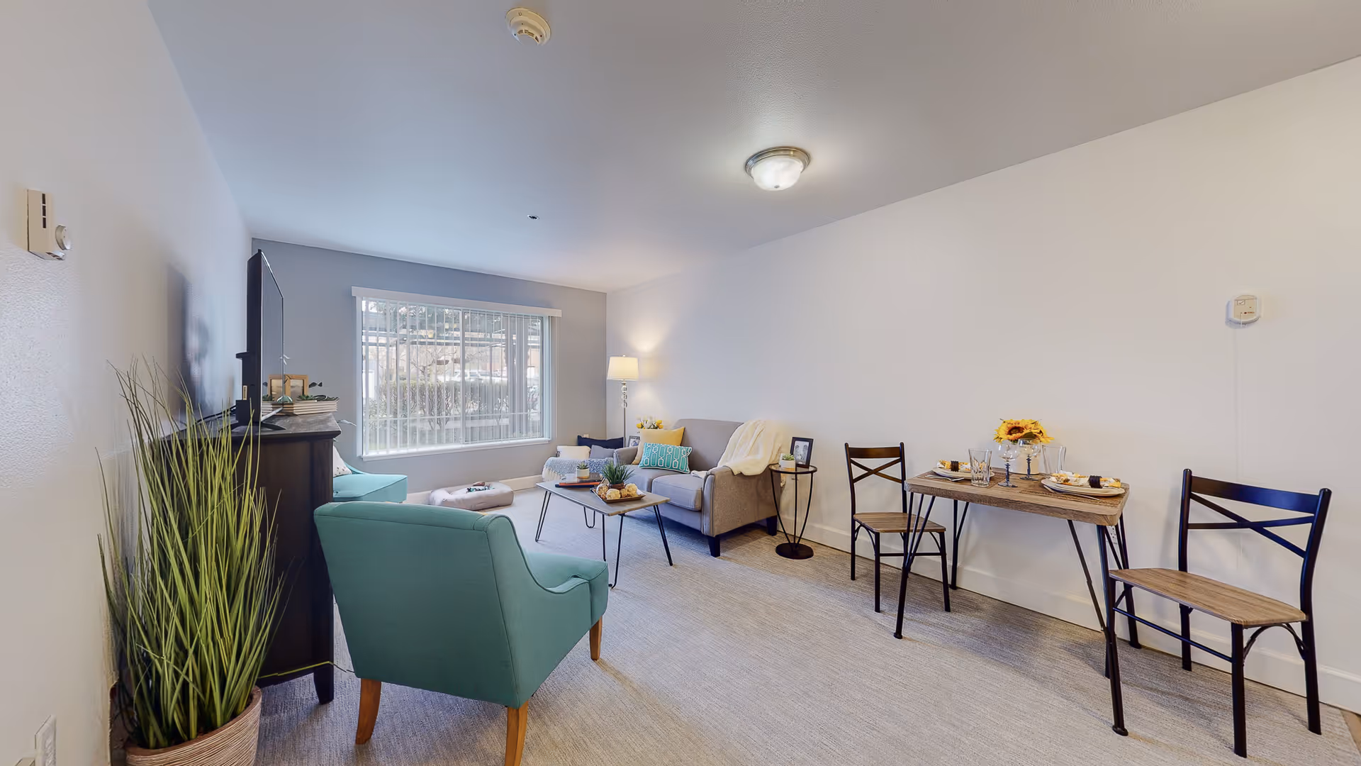 Bright furnished living room with a teal armchair, sofa, coffee table, and a small dining table by a large window.