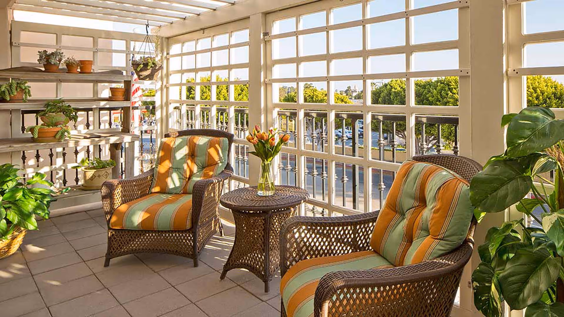 A sunlit enclosed patio area with two wicker chairs featuring striped orange and green cushions, a small wicker table with a vase of tulips, and several potted plants on shelves and around the space. The patio has large windows with a view of trees and a parking lot outside.