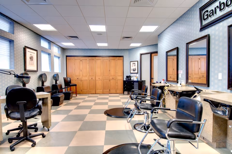Empty salon-style room with multiple styling chairs, mirrors, sinks, and hair-drying stations.