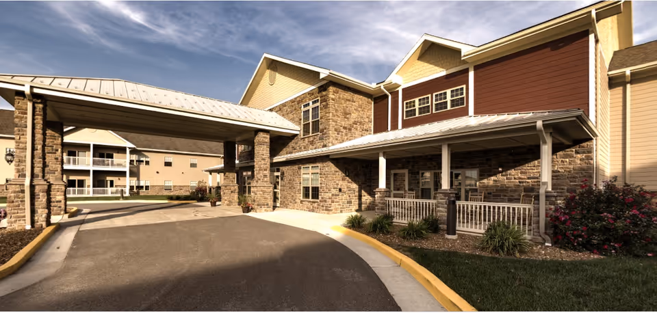 Exterior view of a senior living facility building with stone and siding facade, a covered driveway entrance, landscaped greenery, and a clear sky above.