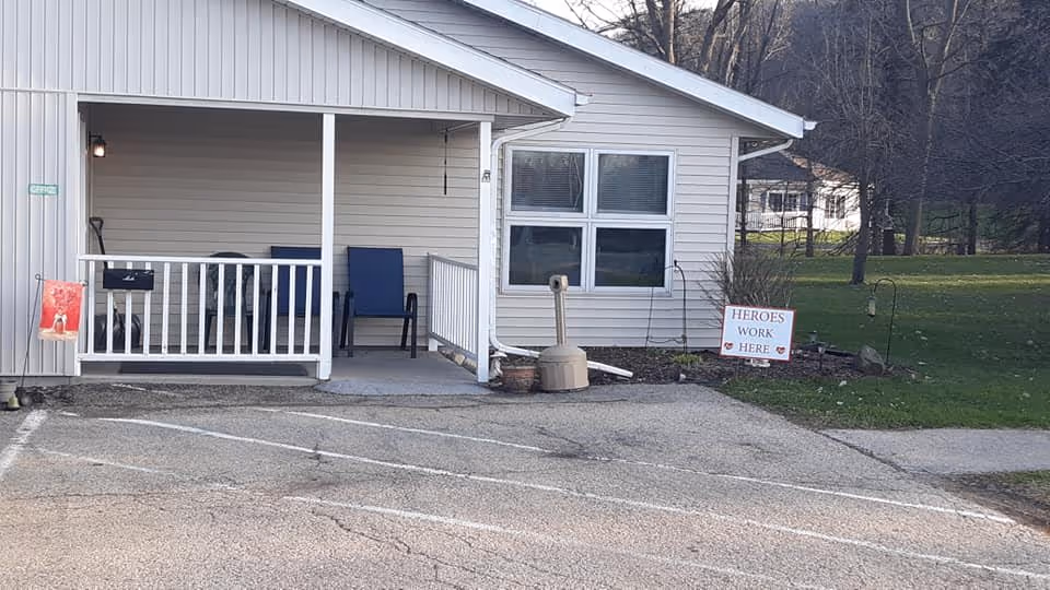Exterior view of a single-story building with beige siding and a small covered porch. The porch has a white railing and two blue chairs. There is a sign on the lawn that reads 'HEROES WORK HERE'. The area around the building includes a paved parking lot and some grass with trees in the background.