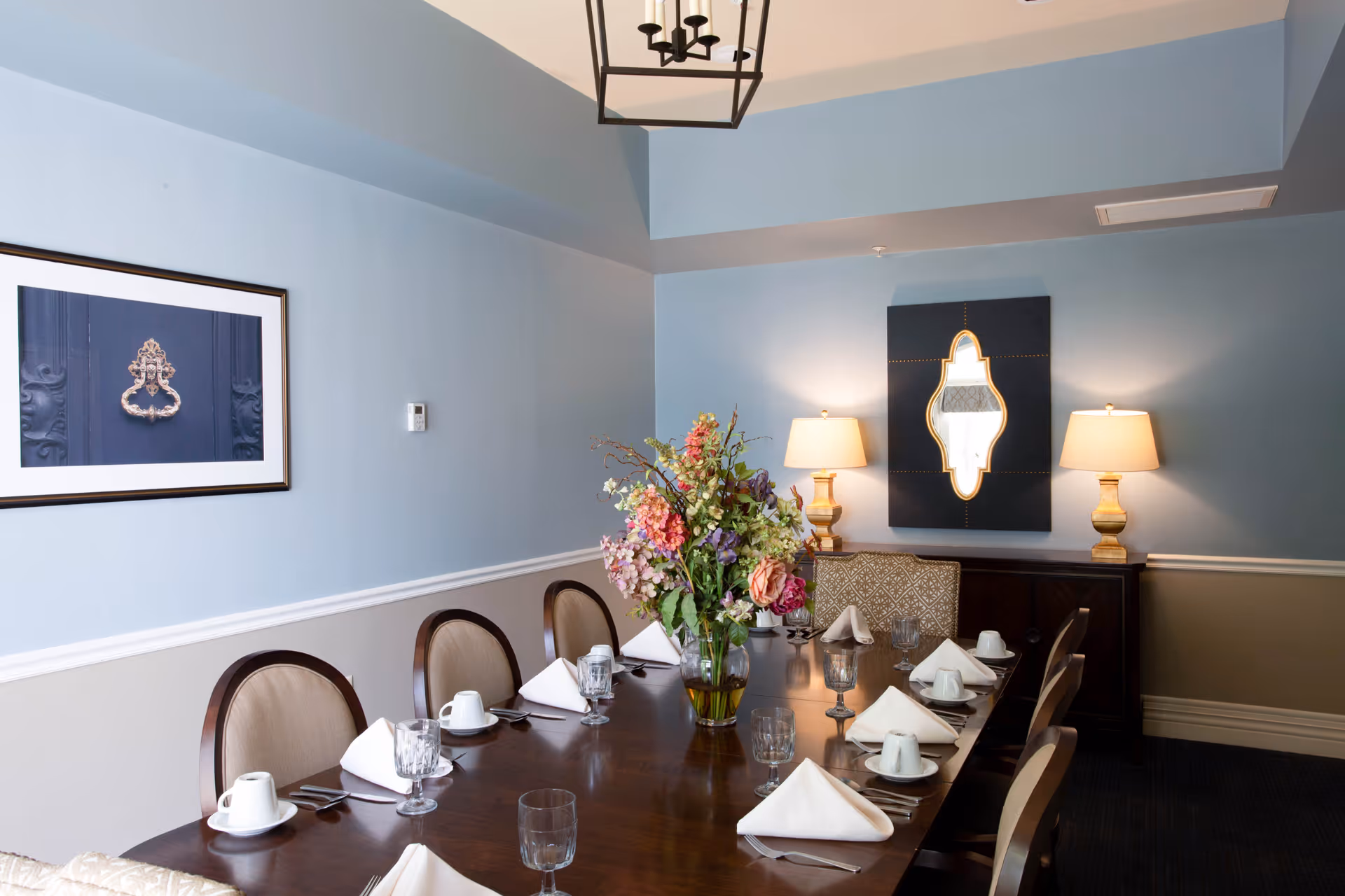 A formal dining room with a long wooden table set for eight people. Each place setting includes a white napkin, a cup and saucer, a glass, and silverware. A large floral arrangement is centered on the table. The walls are painted light blue with white trim, and there is framed artwork on one wall and a decorative mirror flanked by two table lamps on a sideboard. A modern chandelier hangs from the ceiling.