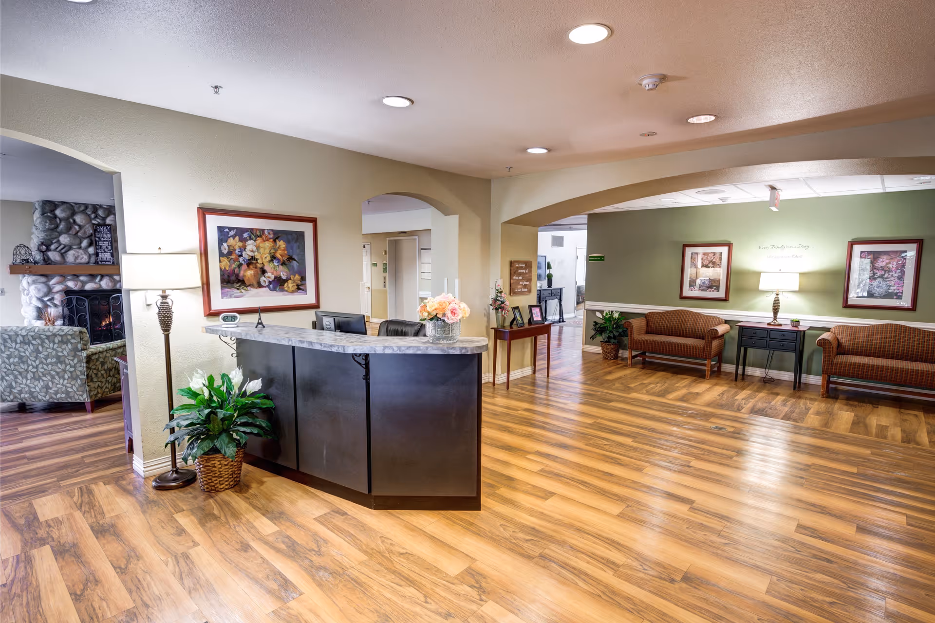 Reception area of The Auberge at North Ogden with a curved black reception desk topped with a marble surface, a basket with green plants beside it, and a floor lamp. The room has wooden flooring, beige walls, and framed floral artwork. In the background, there is a seating area with two brown cushioned benches, a black side table with a lamp, and more framed pictures on a green accent wall. An archway leads to another room with a stone fireplace and patterned armchairs.