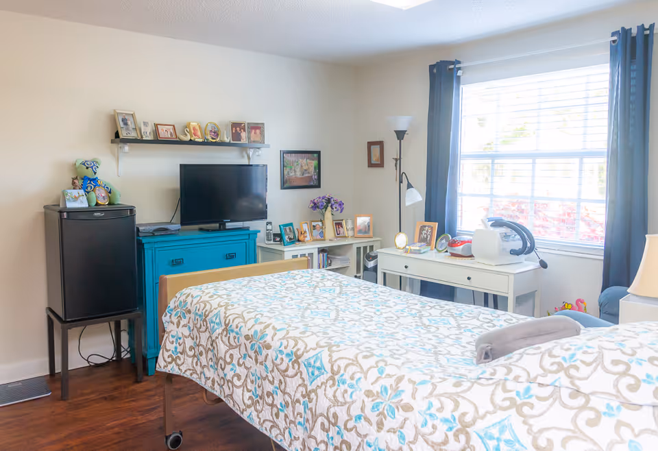 A bright bedroom with a bed covered in a patterned quilt in the foreground. The room features a window with dark curtains, a white desk with various framed photos and a medical device, a blue cabinet with a TV on top, and a small black refrigerator. There are family photos and decorative items on shelves and surfaces, and a floor lamp near the window.