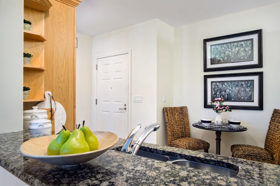 A kitchen area with a granite countertop featuring a bowl of green pears and a modern faucet. In the background, there is a small round dining table set for two with woven chairs, a vase with pink flowers, and two framed floral paintings on the wall.