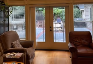 A cozy living room with two armchairs, one brown fabric and one dark brown leather, positioned on either side of a set of glass double doors leading to an outdoor deck area. The room has wooden flooring and windows with white blinds on either side of the doors.
