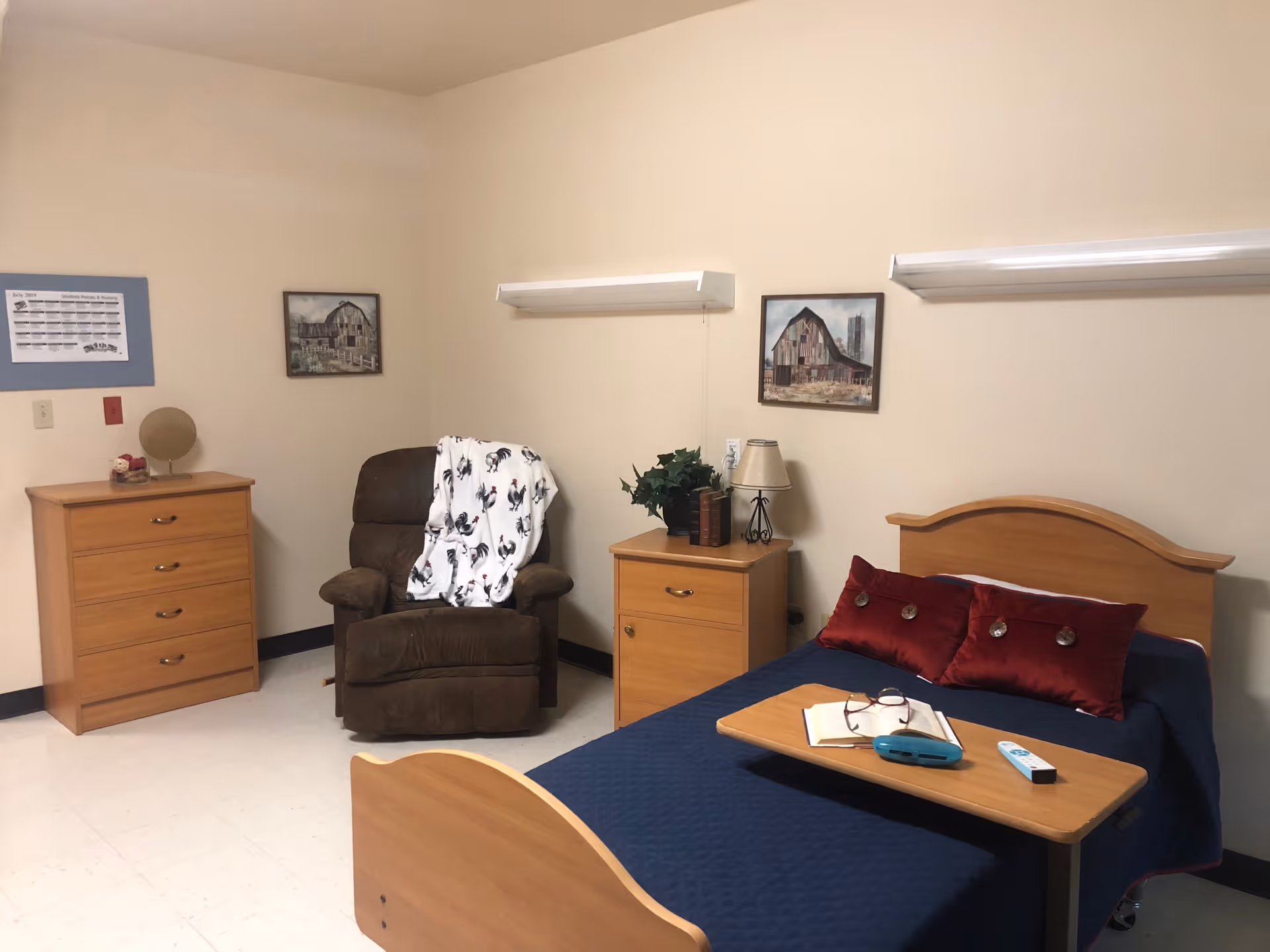 A neatly furnished senior facility bedroom with a single bed, bedside table, recliner, dresser, and wall art.