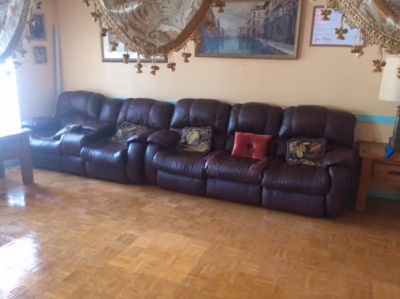 A living room with a long dark brown leather sectional sofa adorned with several decorative pillows, including one red pillow. The room has parquet flooring, a wooden side table with a lamp, and framed artwork on the beige walls. A decorative curtain with tassels partially hangs at the top of the image.