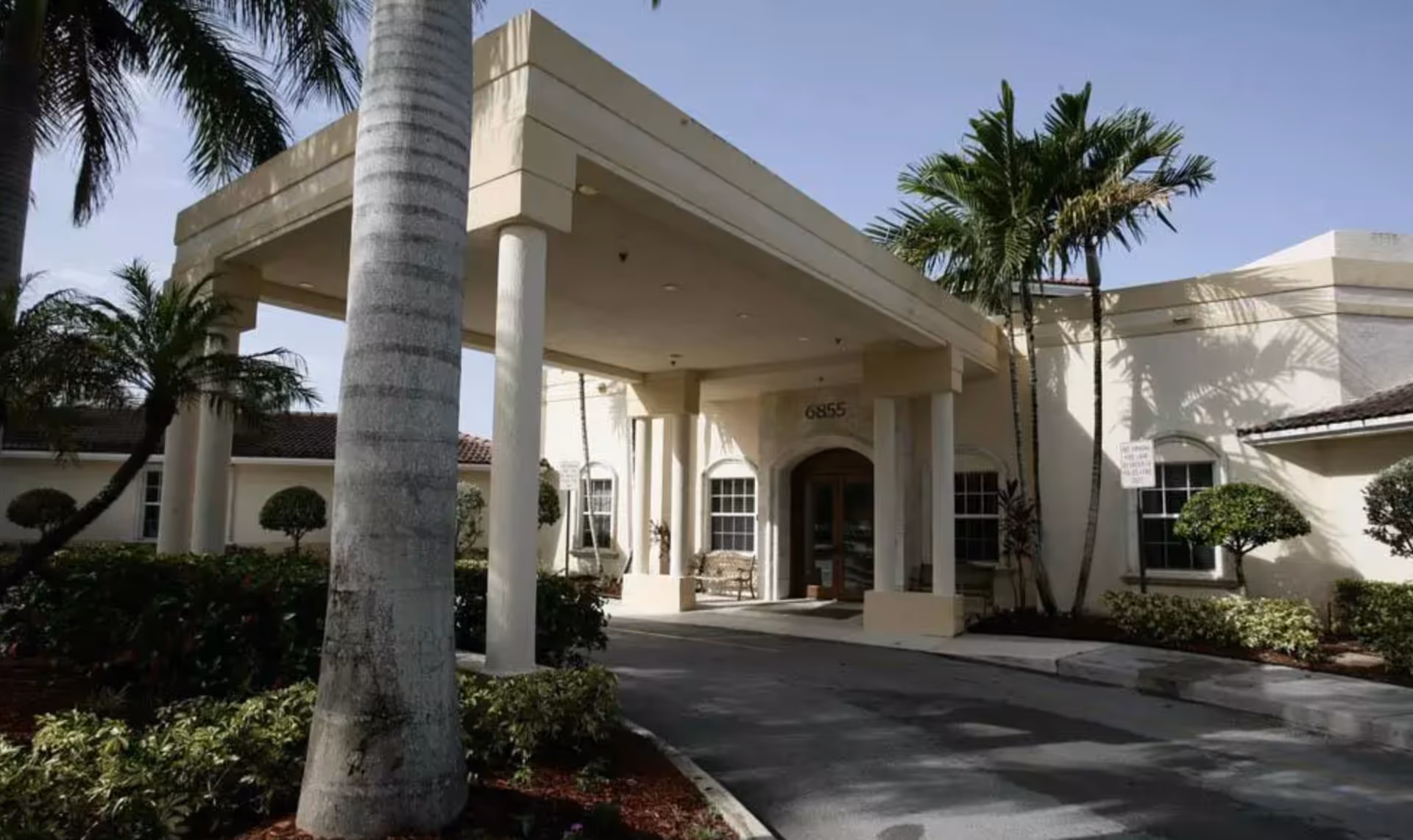 Entrance of a senior living facility with a covered driveway supported by columns, surrounded by palm trees and landscaped bushes under a clear sky.