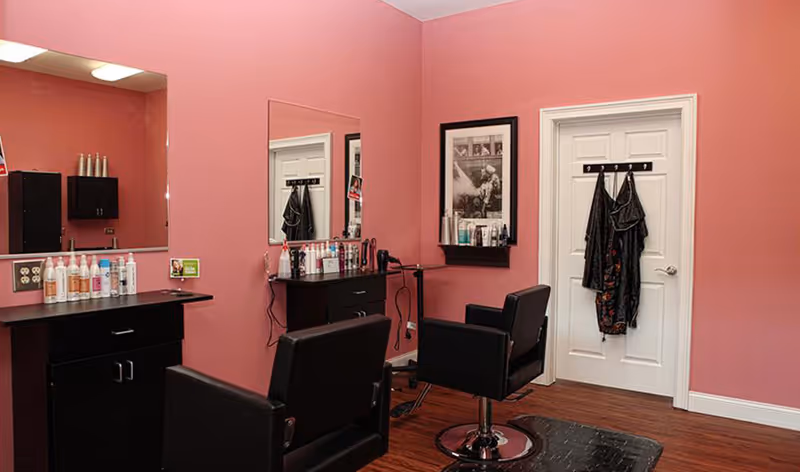 Interior of a salon area with pink walls, two black salon chairs, large mirrors, a counter with hair care products, and a white door with hooks holding black aprons.