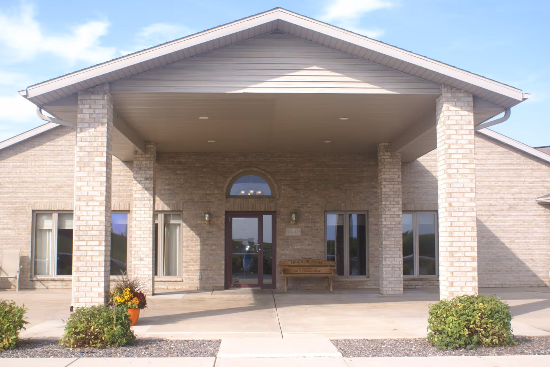 Front exterior view of a single-story brick building with a covered entrance supported by four brick columns. There is a wooden bench and some potted plants near the entrance, and windows on either side of the door.