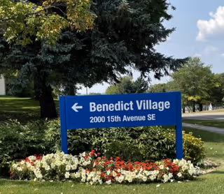 Blue sign for Benedict Village at 2000 15th Avenue SE surrounded by green bushes and red and white flowers, with trees and a road in the background under a partly cloudy sky.