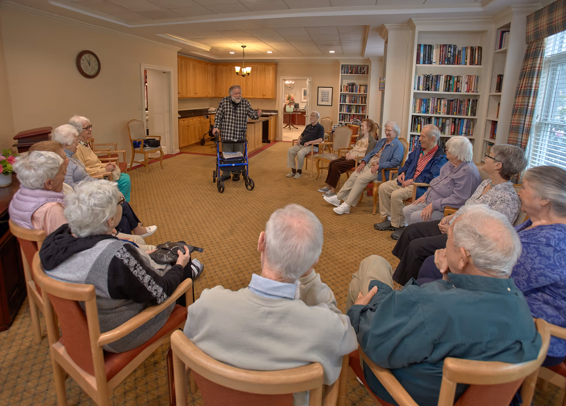 A group of elderly residents seated in a circle in a common room while a man with a walker speaks to them.
