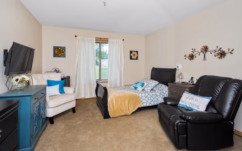 A cozy bedroom in an assisted living facility featuring a single bed with blue and white bedding and a yellow blanket, a black leather recliner with a pillow that says 'Life is Beautiful', a beige armchair with blue and white pillows, a blue cabinet with a vase of flowers, a wall-mounted TV, and a window with white curtains letting in natural light.