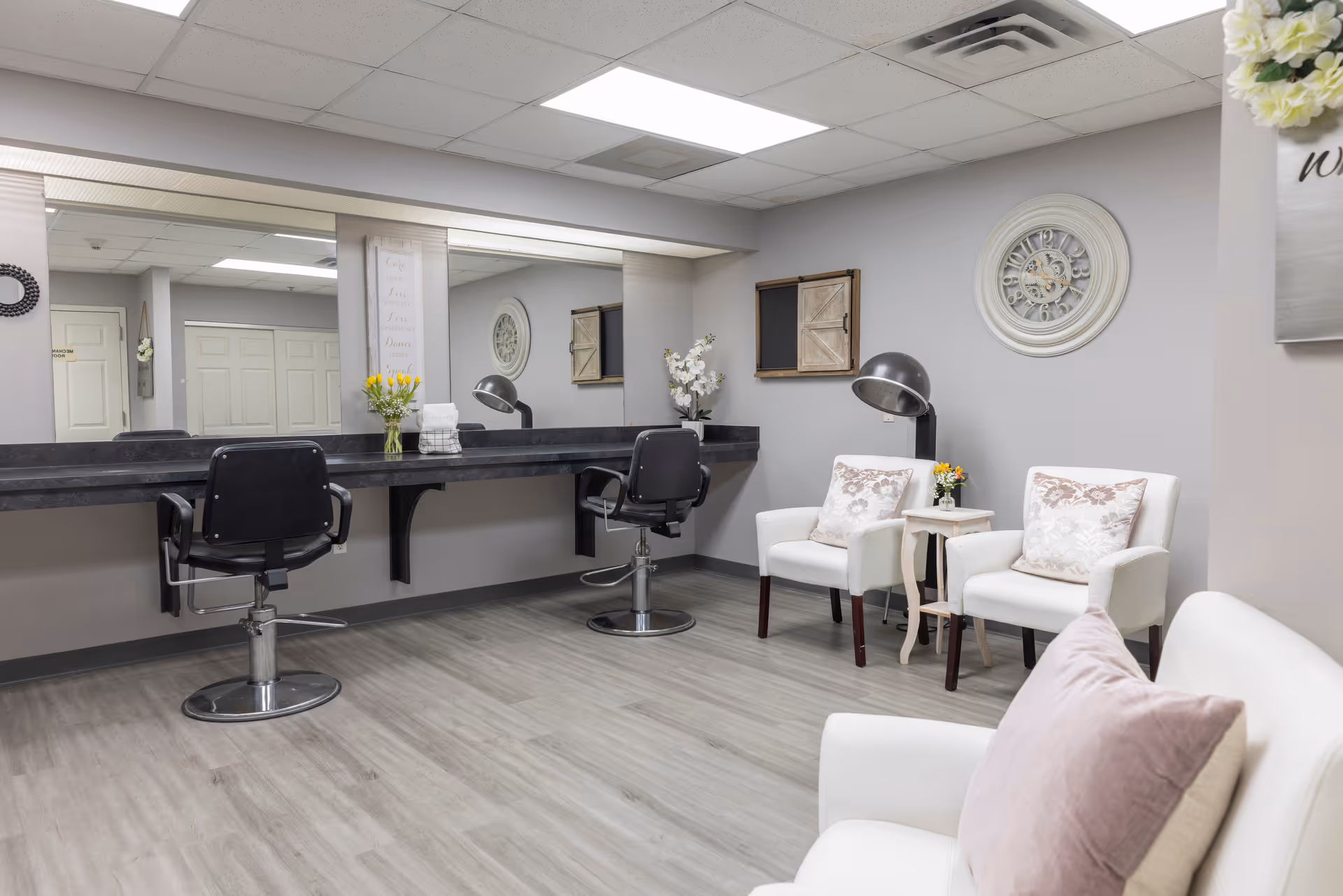 A bright and clean salon area with two black salon chairs in front of a long mirror mounted on the wall. To the right, there are two white armchairs with decorative pillows and a small side table with flowers. The walls are light gray with a large decorative clock and framed wall art. The floor is light wood, and the ceiling has recessed lighting.