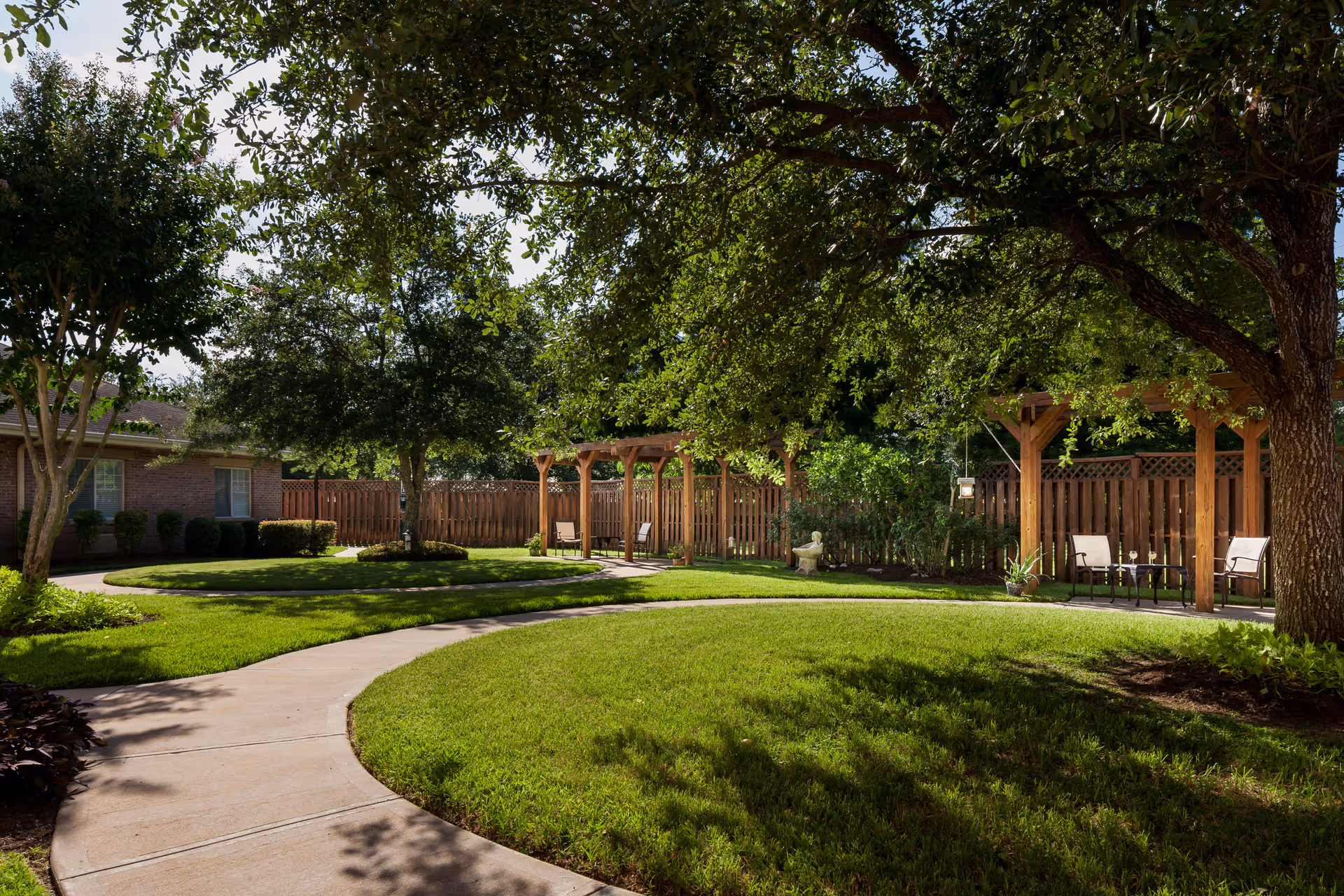 A peaceful outdoor garden area with a curved concrete pathway surrounded by green grass and trees. There are wooden pergolas with chairs and small tables underneath, and a wooden fence enclosing the space. The area is shaded by large trees, creating a serene and inviting atmosphere.