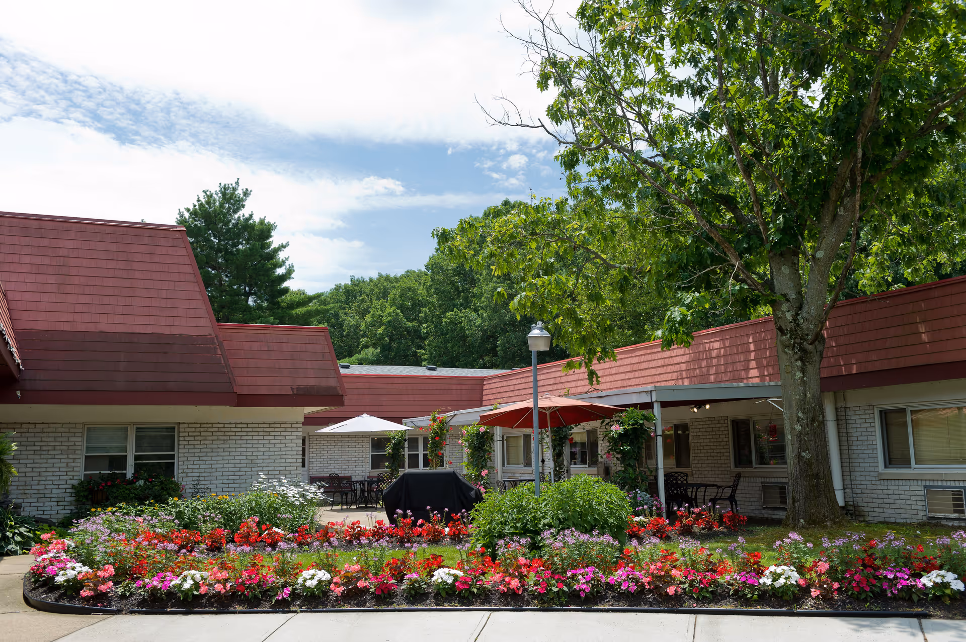 Outdoor courtyard area of a senior living facility with a variety of colorful flowers planted in a garden bed in the foreground. The building has white brick walls and red sloped roofs. There are patio tables with umbrellas and chairs under a covered area, and a large tree providing shade. The sky is partly cloudy with blue patches visible.