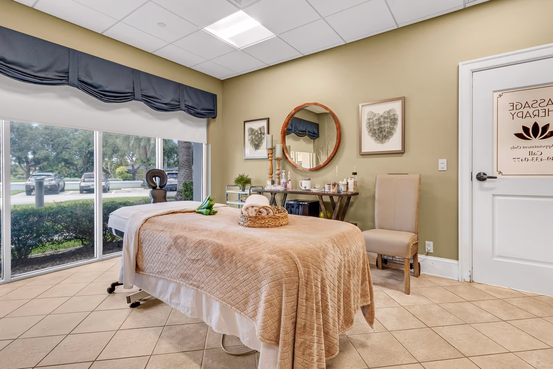 A massage therapy room with a treatment table covered in a beige quilt, large window, round mirror, chair, and a door labeled "Massage Therapy".