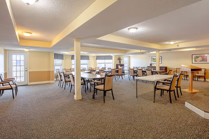 A spacious dining room in Elison Assisted Living of Oxford with multiple tables and chairs arranged neatly. The room has beige walls, carpeted floors, ceiling lights, and several windows with blinds allowing natural light to enter. There is a wooden podium on the right side and framed pictures on the walls.