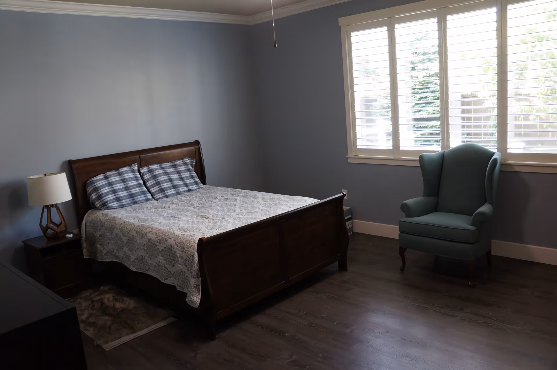 Bedroom with a wooden bed, patterned bedspread, nightstand and lamp, and a green wingback chair by shuttered windows.