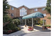 Front entrance of a three-story brick senior living building with a green canopy, driveway, and landscaped flowerbeds.