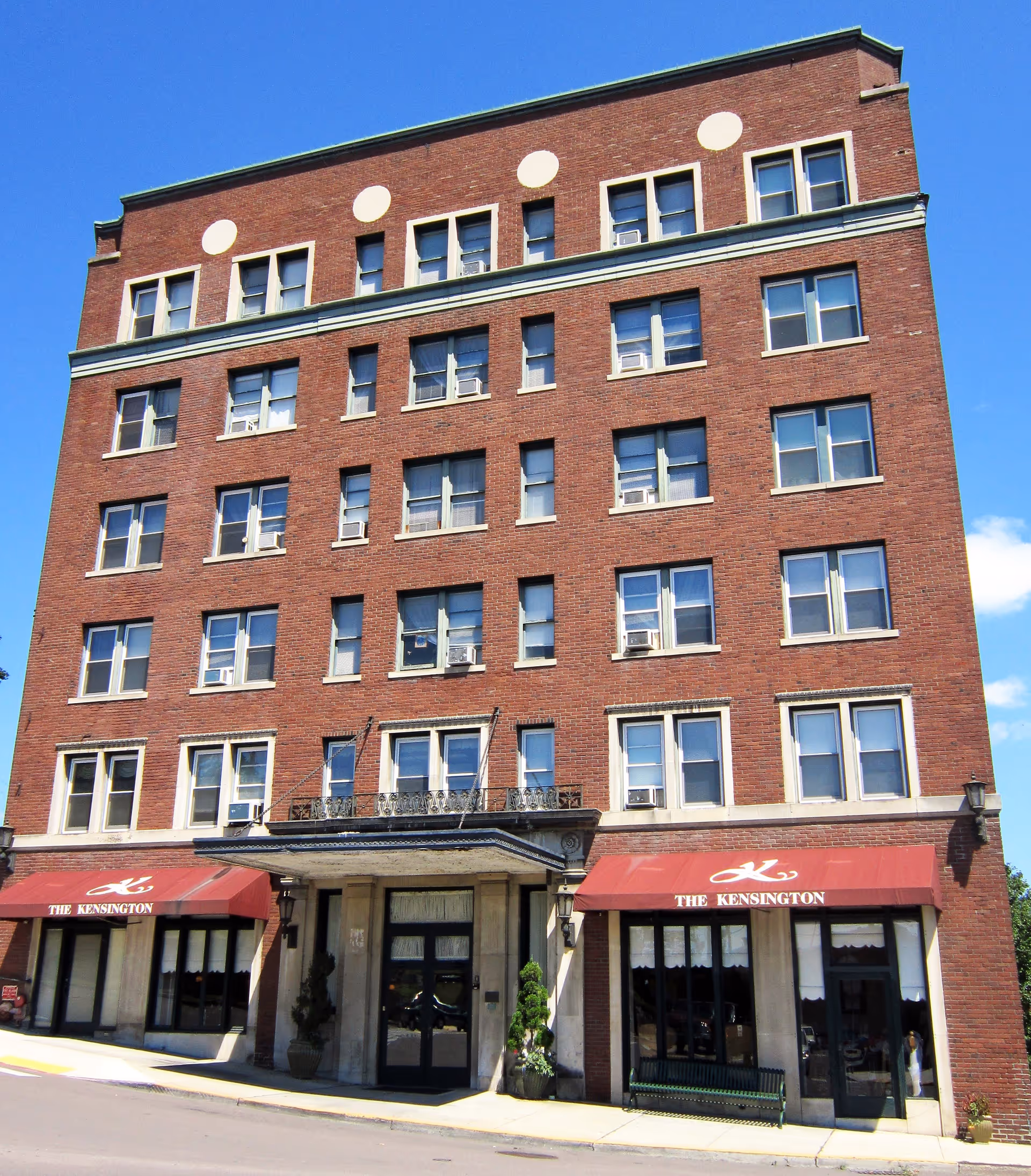 Exterior view of a multi-story brick building with many windows and two red awnings labeled 'The Kensington' above the entrance doors, under a clear blue sky.