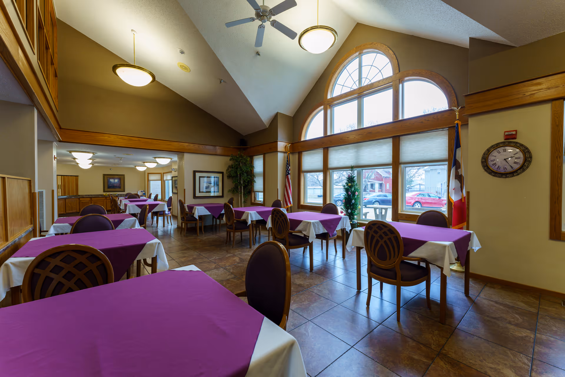 Interior view of a dining room with multiple tables covered in white tablecloths and purple overlays. The room features large arched windows letting in natural light, a ceiling fan, and a clock on the wall. There are chairs around the tables and flags near the windows.