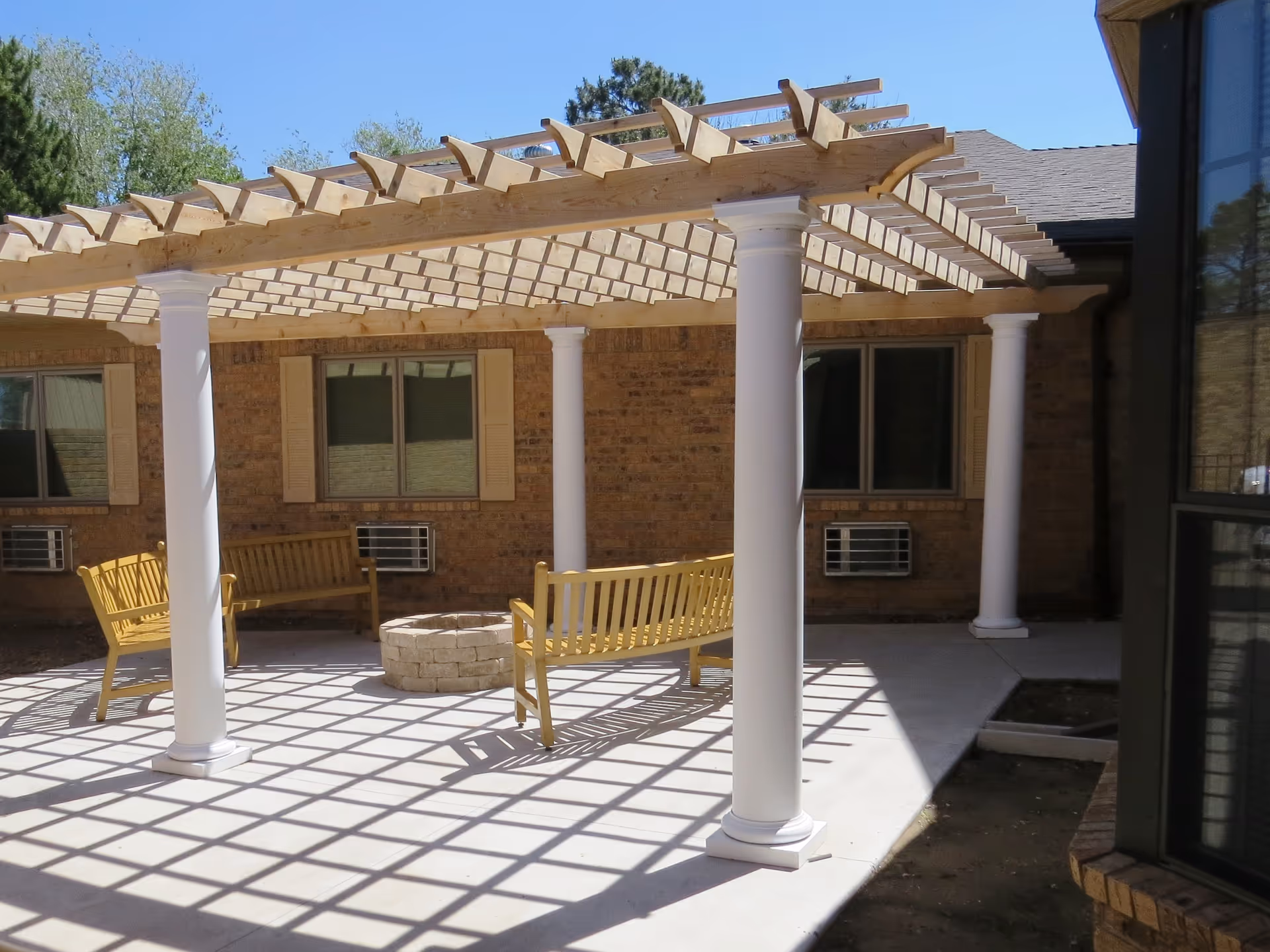 Outdoor patio area with a wooden pergola supported by white columns casting shadows on the concrete floor. There are wooden benches arranged around a circular stone fire pit. The background shows a brick building with windows and air conditioning units.