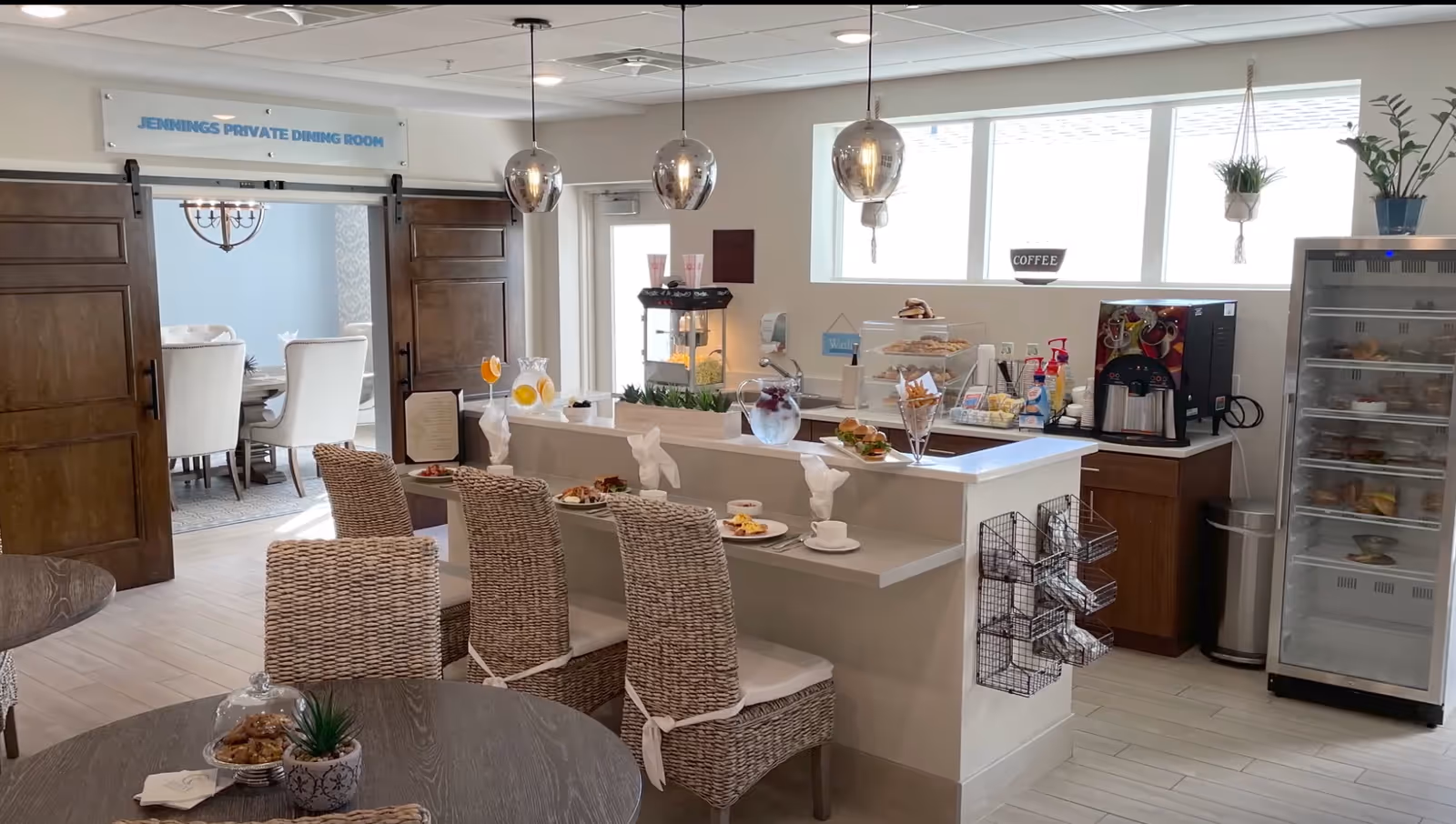 Bright senior living dining area with a breakfast bar and woven chairs, coffee station, refrigerated display, and an adjoining private dining room.