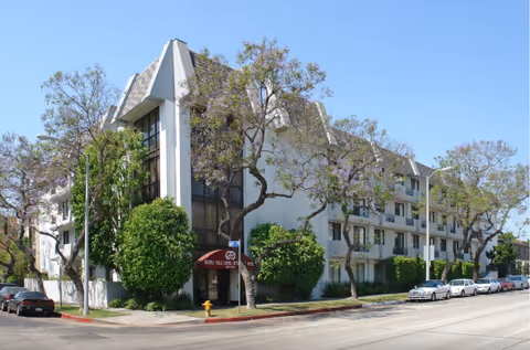 Exterior view of a multi-story residential building with balconies, surrounded by trees and parked cars along the street under a clear blue sky.