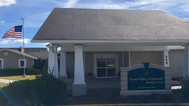 Front exterior view of Green Valley Commons Assisted Living facility with a covered entrance, white pillars, an American flag on a flagpole to the left, and a sign displaying the facility name and address number 549.
