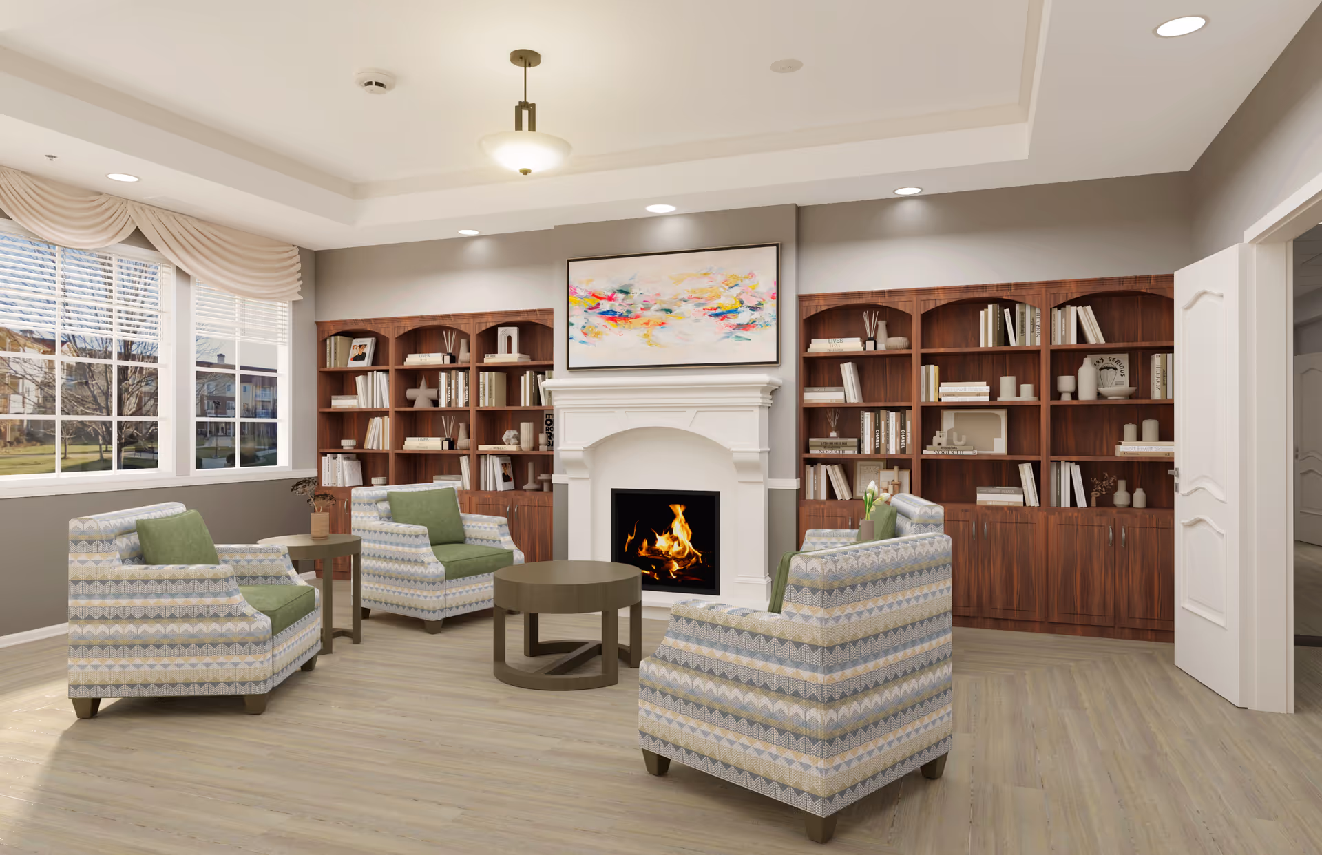 A cozy senior living room with four patterned armchairs featuring green cushions arranged around a round coffee table in front of a white fireplace with a colorful abstract painting above it. Behind the fireplace are two large wooden bookshelves filled with books and decorative items. Large windows with beige curtains let in natural light, and the room has light wood flooring and recessed ceiling lights.