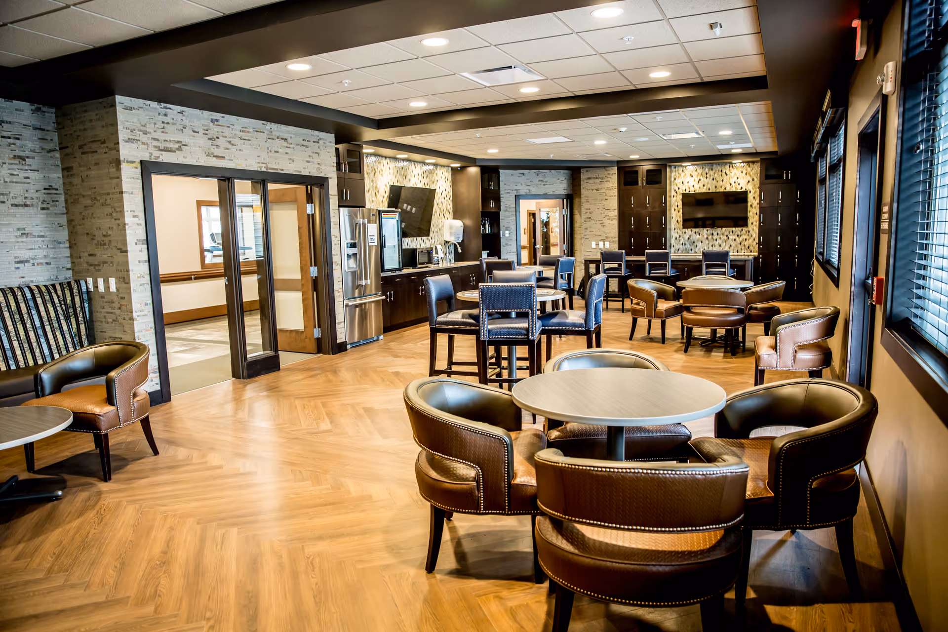 A spacious and well-lit common area with multiple round tables surrounded by leather chairs. The room features a wooden floor with a herringbone pattern, large windows with blinds on the right, and a kitchenette area with stainless steel appliances and a mounted TV in the background. The walls have a modern stone and tile design, and there are additional seating areas with high chairs and armchairs.