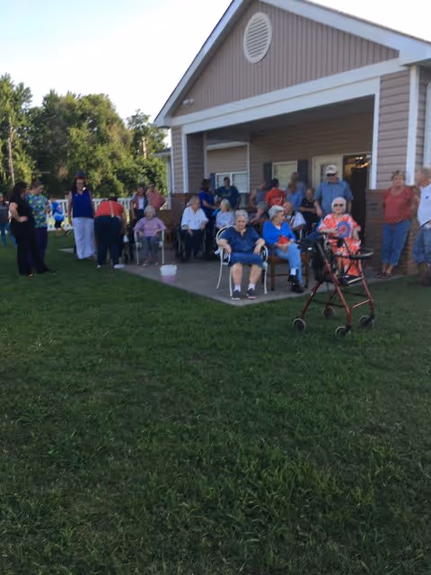 Several elderly residents and staff gathered on a patio and lawn in front of a single-story assisted-living building.