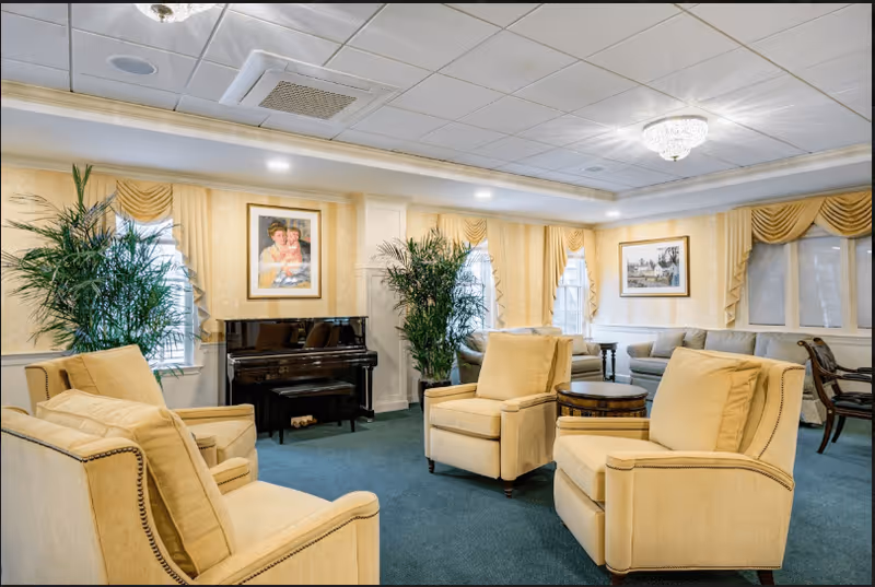 A cozy living room area with four beige armchairs arranged around a small round wooden table. The room has green carpet flooring, cream-colored walls with framed paintings, large windows with yellow drapes, and two tall green plants. A black upright piano with a bench is positioned against one wall. The ceiling has recessed lighting and a decorative chandelier.