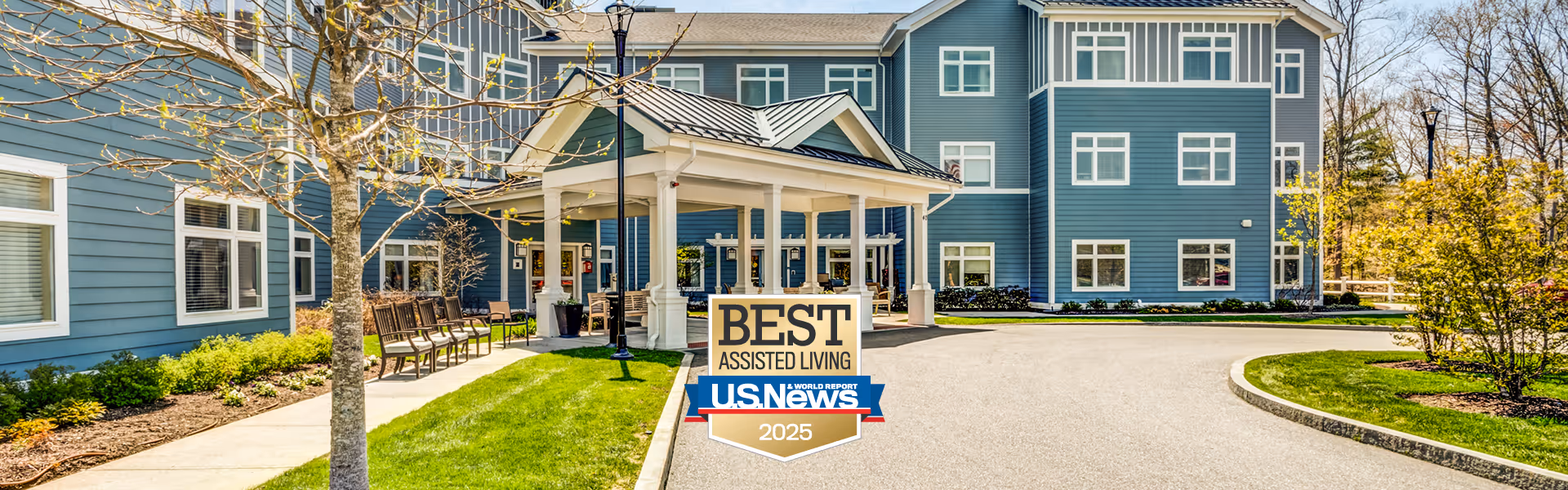 Exterior view of a blue assisted living facility building with multiple windows and a covered entrance. There is a paved driveway and landscaped greenery including trees and bushes around the building. A sign in front reads 'Best Assisted Living U.S. News 2025'.