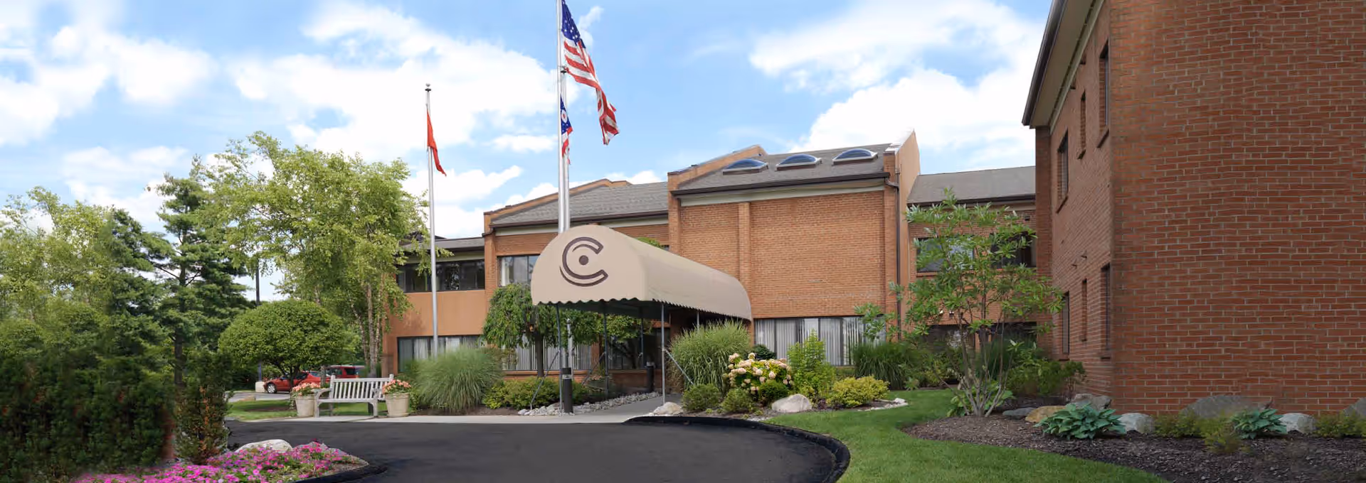 Front entrance of a brick senior living building with a tan canopy, flags, and landscaped driveway.