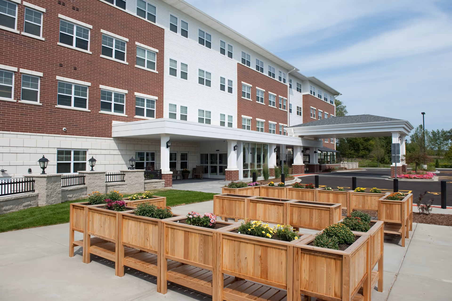 Exterior view of a multi-story senior living facility named Oak Hill with a covered entrance and raised wooden garden beds filled with flowers in the foreground.