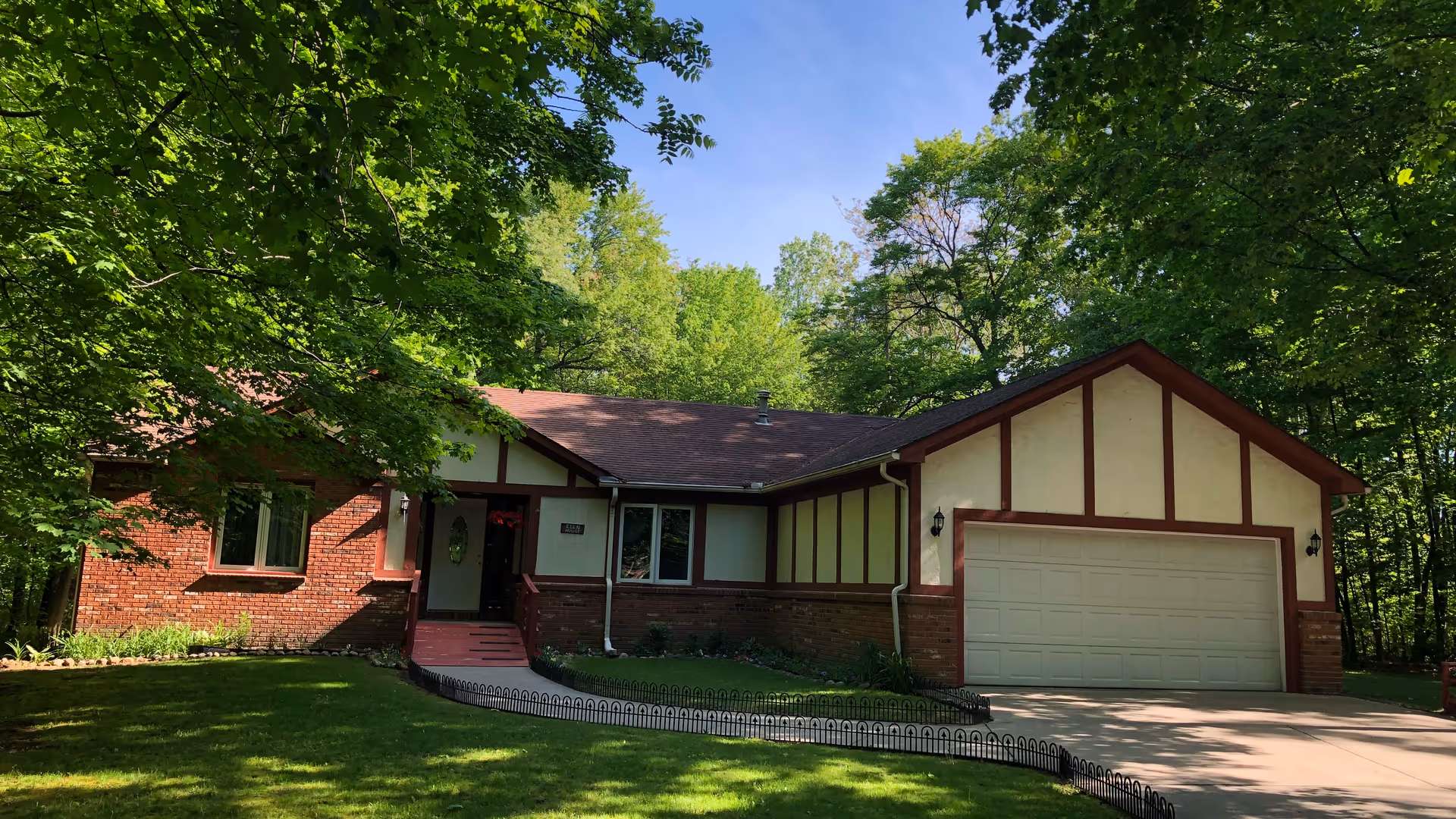 Front exterior view of a single-story house with a brick and beige facade, a two-car garage, surrounded by green trees and a well-maintained lawn under a clear blue sky.