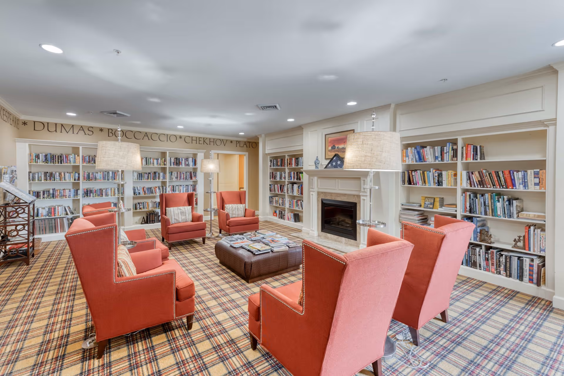 A cozy library or reading room with red upholstered armchairs arranged around a central ottoman covered with magazines. The room features built-in white bookshelves filled with books, a fireplace with a framed picture and a folded American flag on the mantel, and several standing lamps providing warm lighting. The carpet has a plaid pattern, and the walls have names of famous authors written near the ceiling.