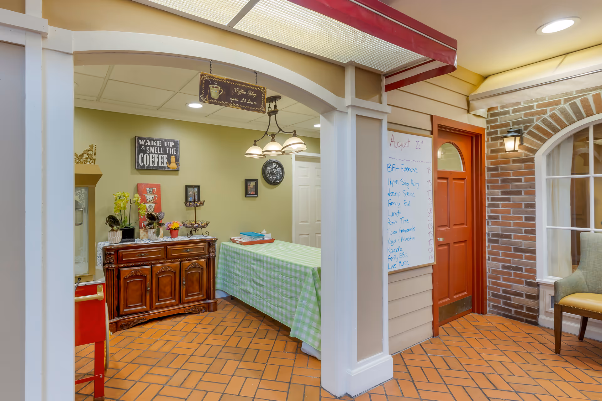 Interior view of a senior living facility area with a checkered green and white tablecloth on a table, a wooden cabinet with decorative items and flowers, a hanging sign that reads 'Coffee Shop open 24 hours,' a wall clock, and a whiteboard with a schedule. There is a red door and a brick wall with a window and a chair nearby.