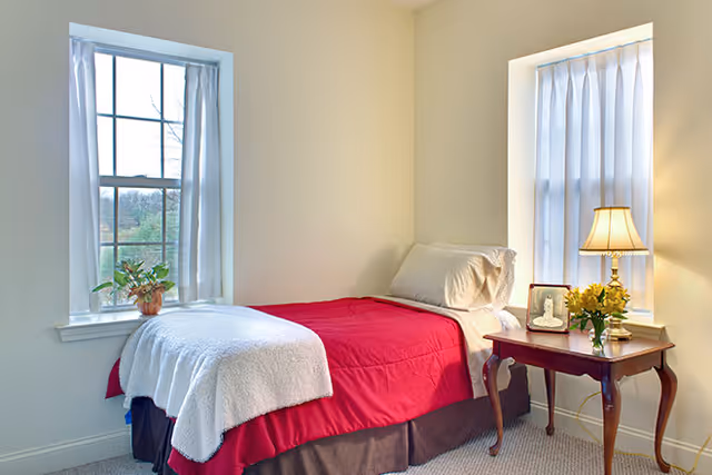 A small bedroom with a single bed covered in a red blanket and white throw, positioned between two windows with sheer white curtains. A wooden side table next to the bed holds a lamp, a framed photo, and a vase of yellow flowers. A potted plant sits on the windowsill to the left.