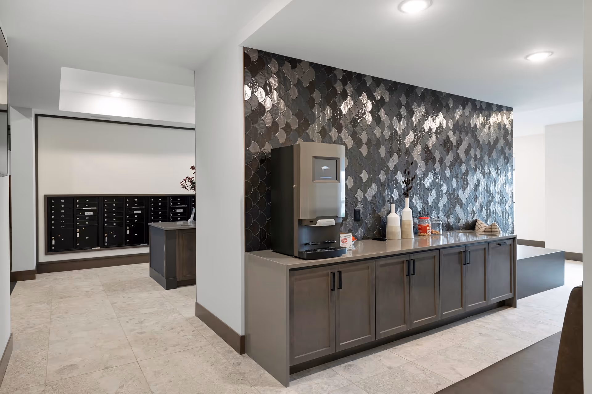 Communal interior with a long cabinet holding a beverage machine and decorative vases against a dark patterned tile wall, with mailboxes visible in the background.
