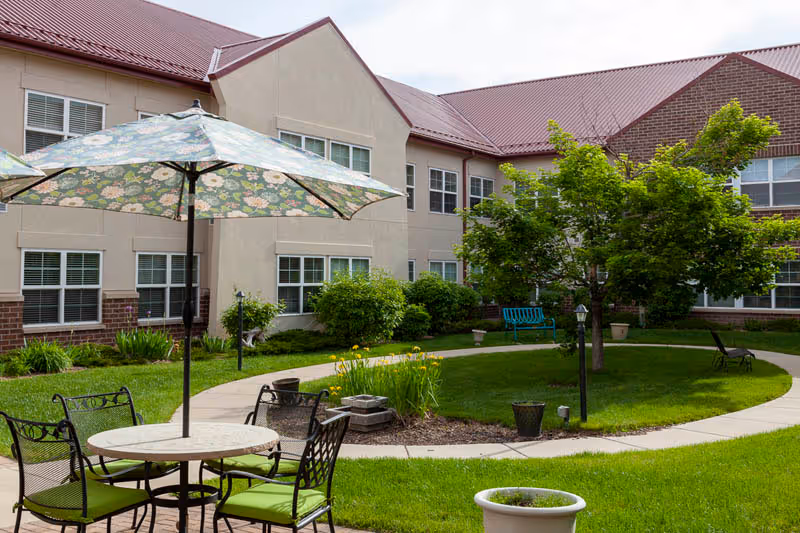 Sunlit courtyard with a patio table and umbrella, green lawn, trees, benches, and a surrounding two-story senior living building.