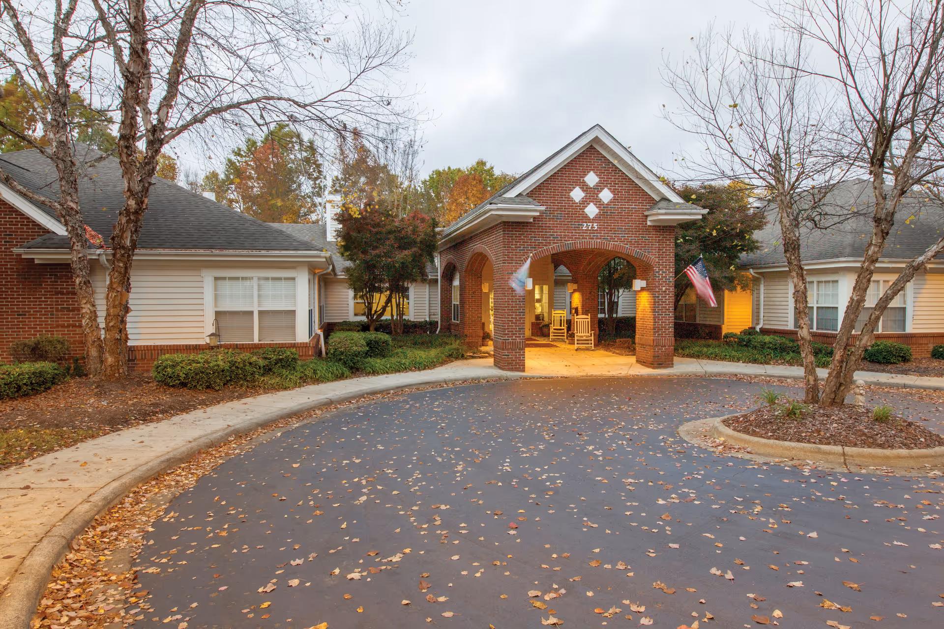 Entrance of a senior living facility with a brick archway and covered driveway. The building has beige siding with brick accents, surrounded by trees with autumn leaves scattered on the ground. Two rocking chairs are visible under the archway, and an American flag is displayed on the right side.